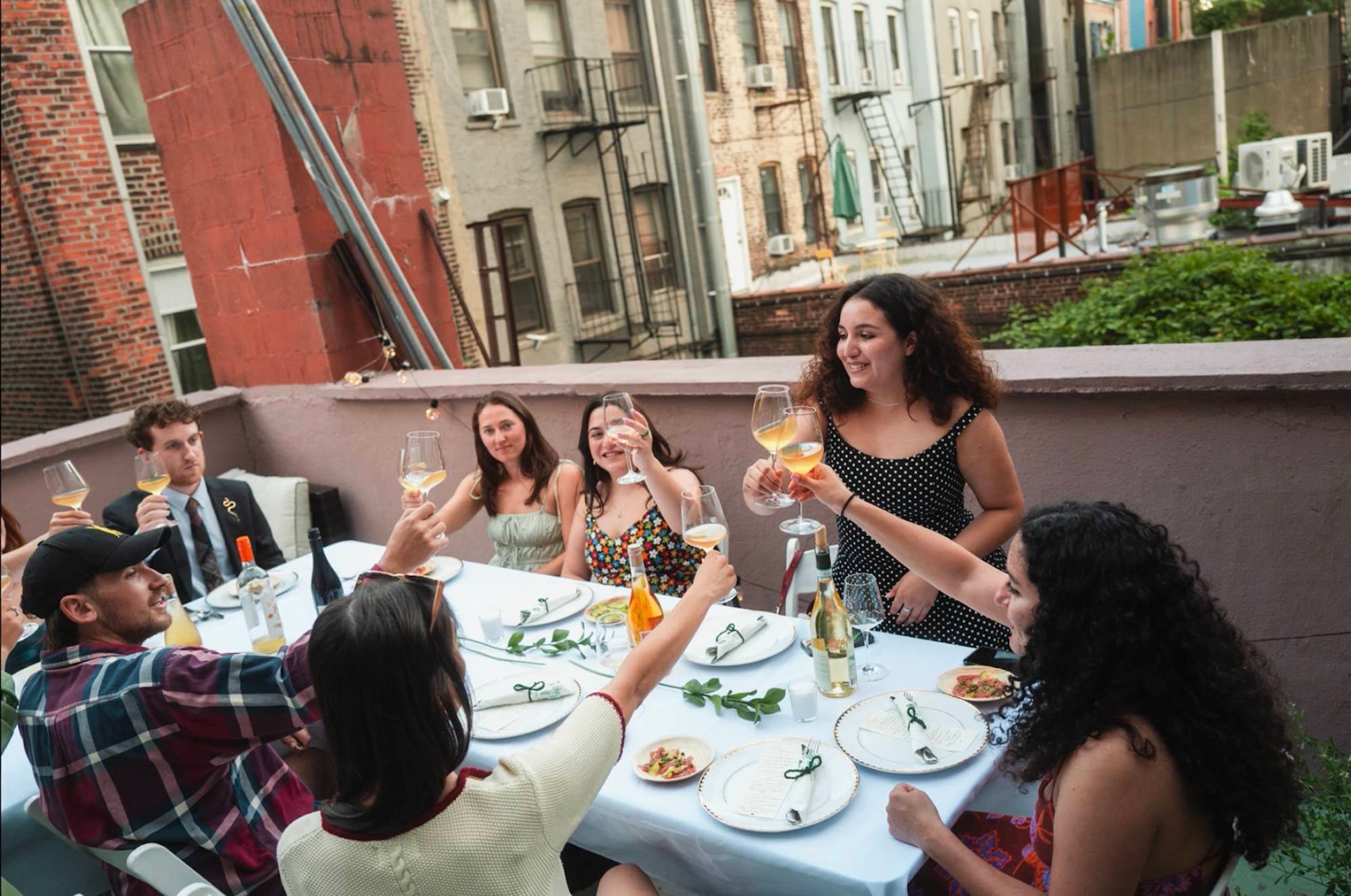 A group of friends sitting around a dinner table outside raise their glasses of white wine for a cheers.