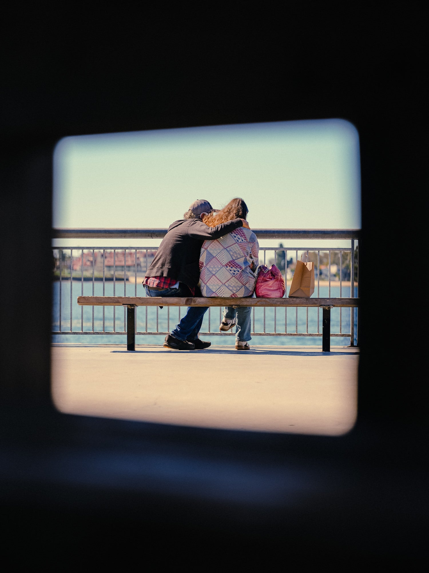 Candid couple sharing a bench on a waterfront promenade, framed through a window, documentary street photo. Candid couple sharing a bench on a waterfront promenade, framed through a window, documentary street photo.