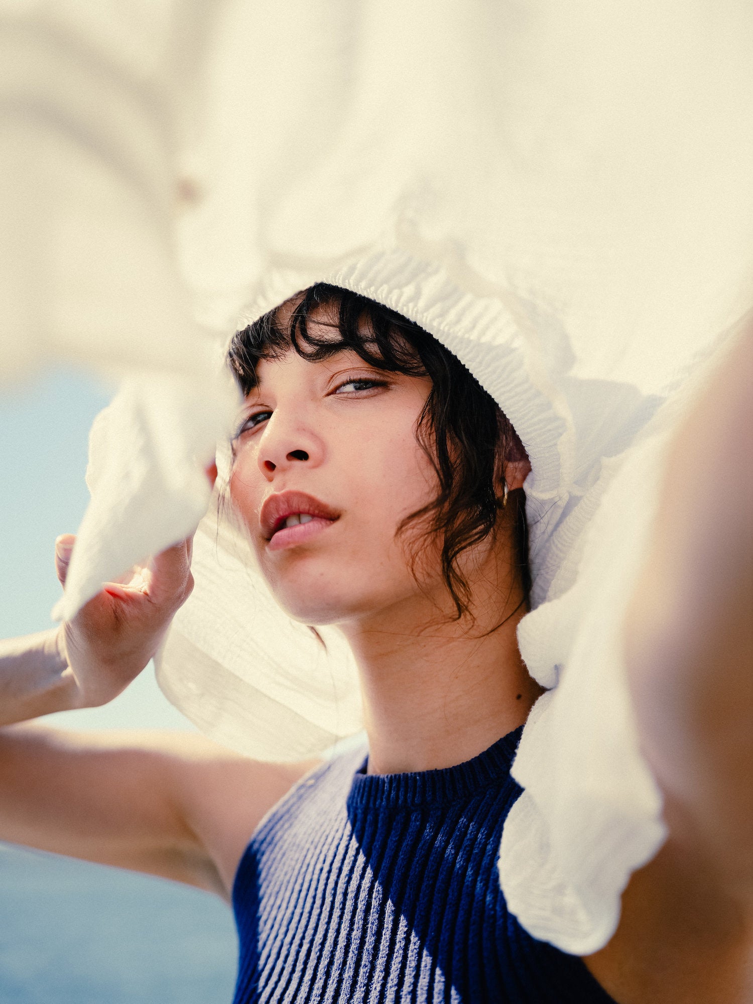 Close‑up portrait framed by a flowing white hat, bright seaside light, editorial beach style. Close‑up portrait framed by a flowing white hat, bright seaside light, editorial beach style.