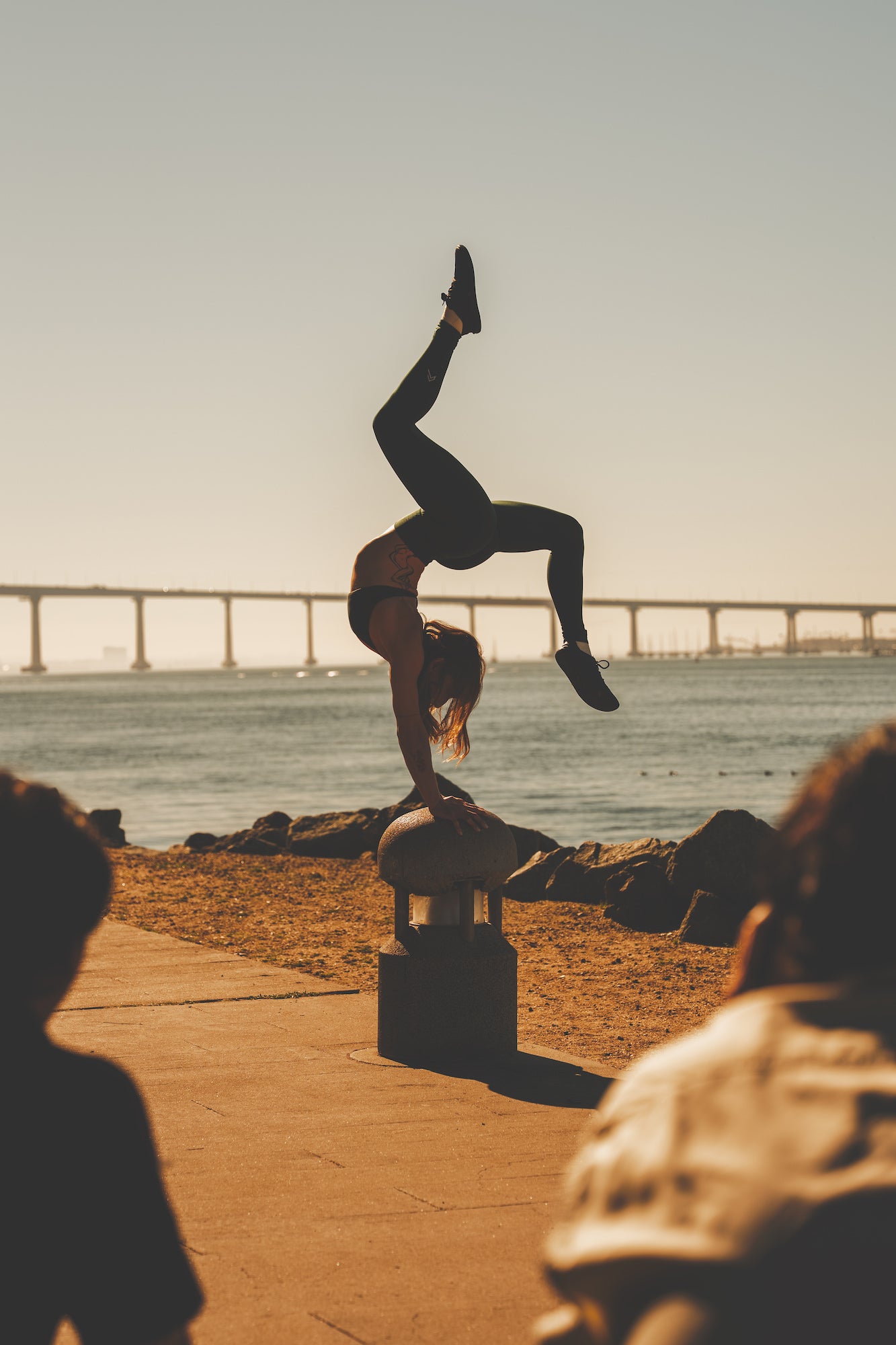 Acrobat performing a one‑hand handstand on a bollard with a long arched bridge over the bay in the background, sunset fitness shot. Acrobat performing a one‑hand handstand on a bollard with a long arched bridge over the bay in the background, sunset fitness shot.