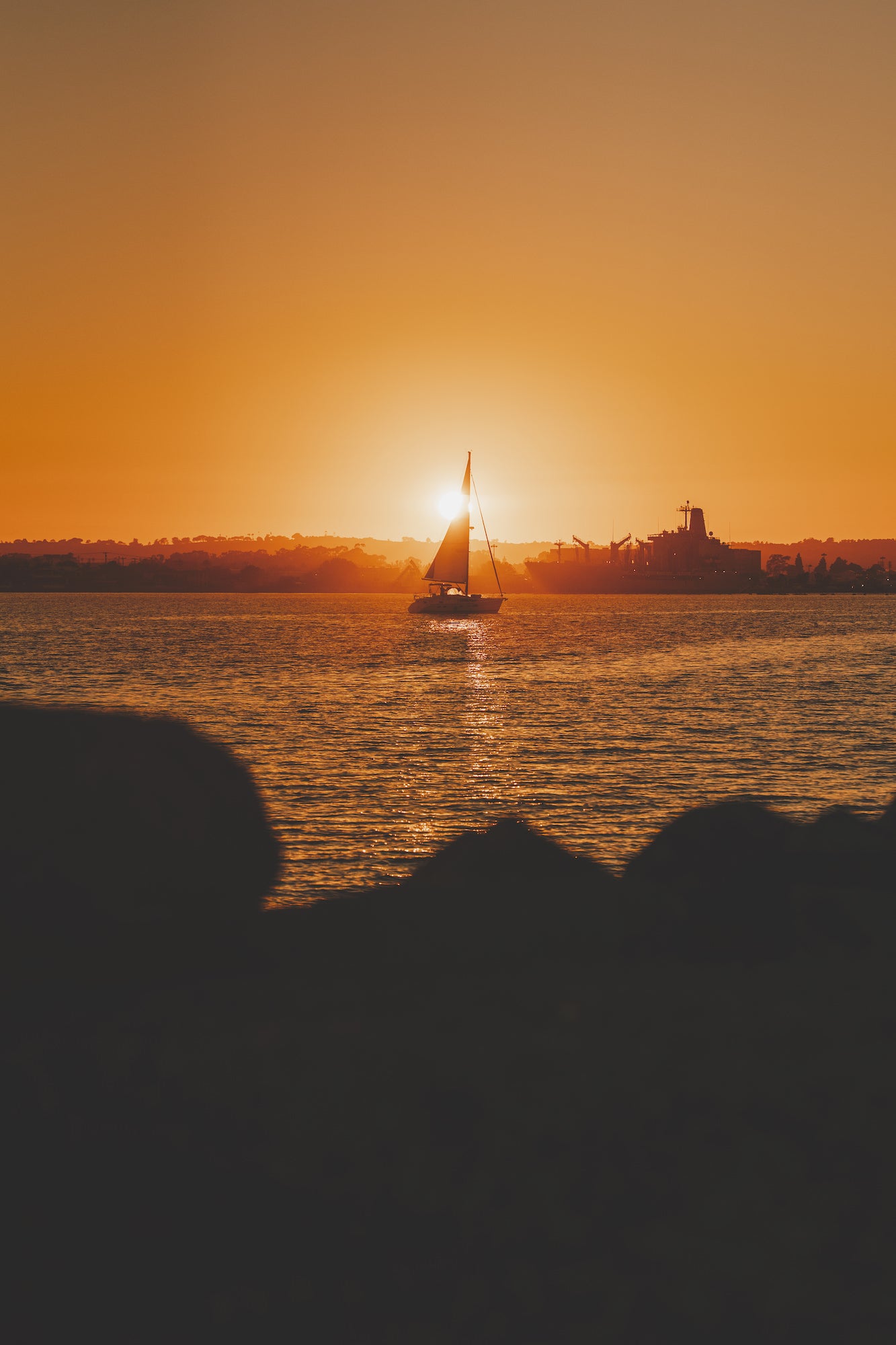 Sailboat silhouette at vivid orange sunset on the bay with distant skyline, San Diego seascape photography. Sailboat silhouette at vivid orange sunset on the bay with distant skyline, San Diego seascape photography.