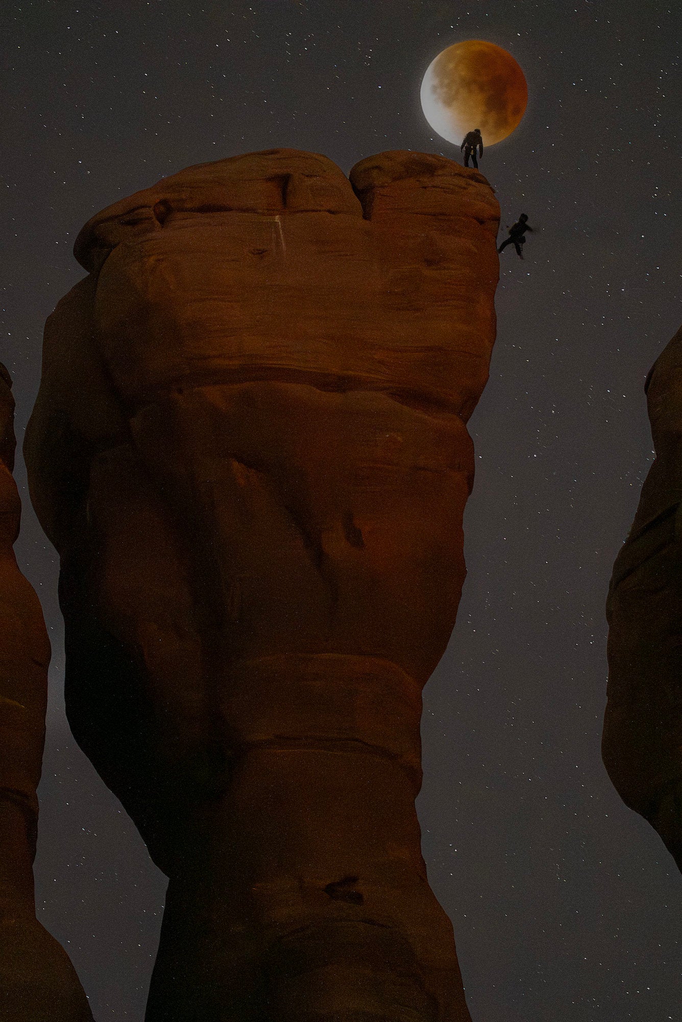 Eclipsed moon above a sandstone pillar as one climber stands and another jumps between towers at night.