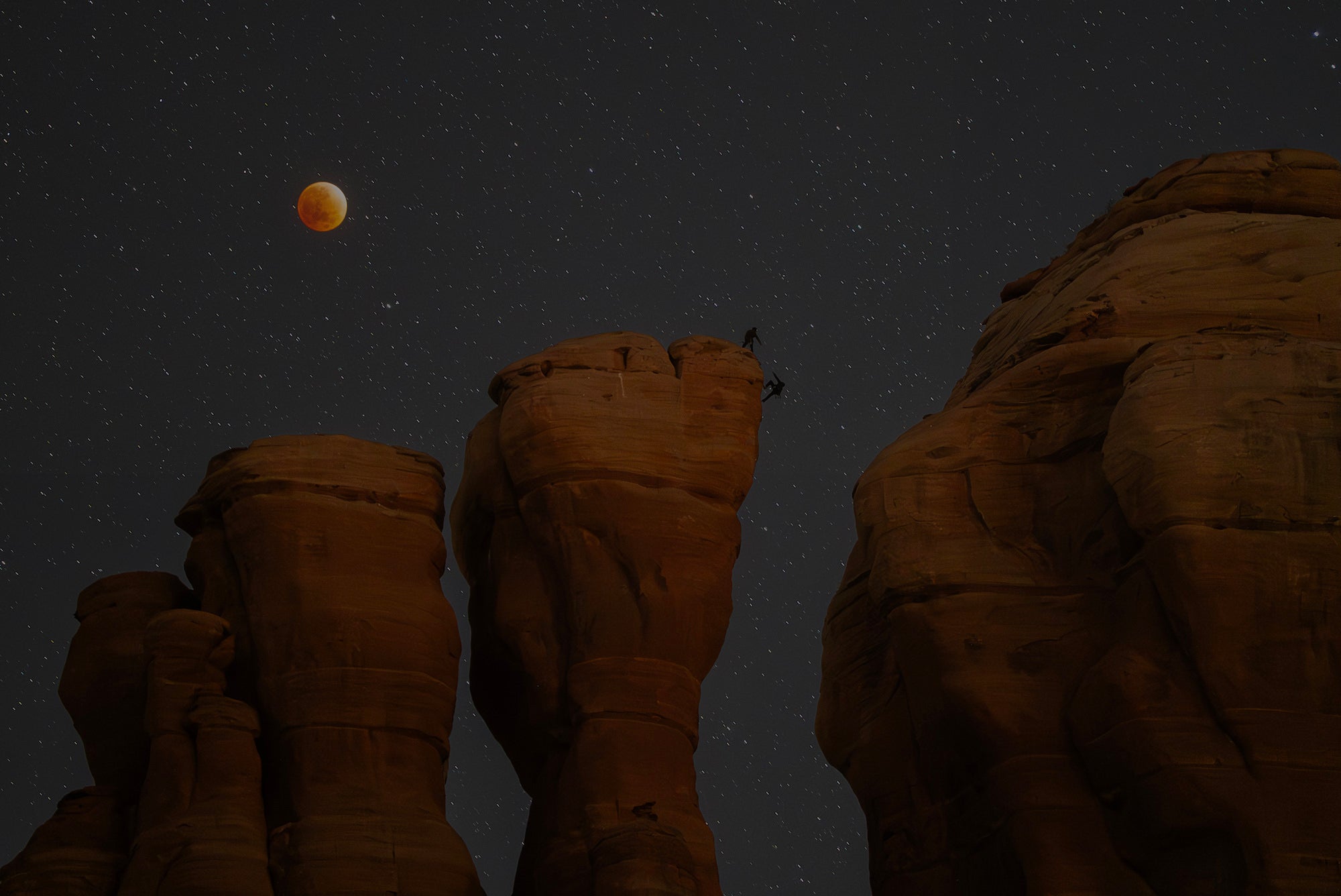 Blood moon over starry sky with tall sandstone towers and two climbers silhouetted on a summit.