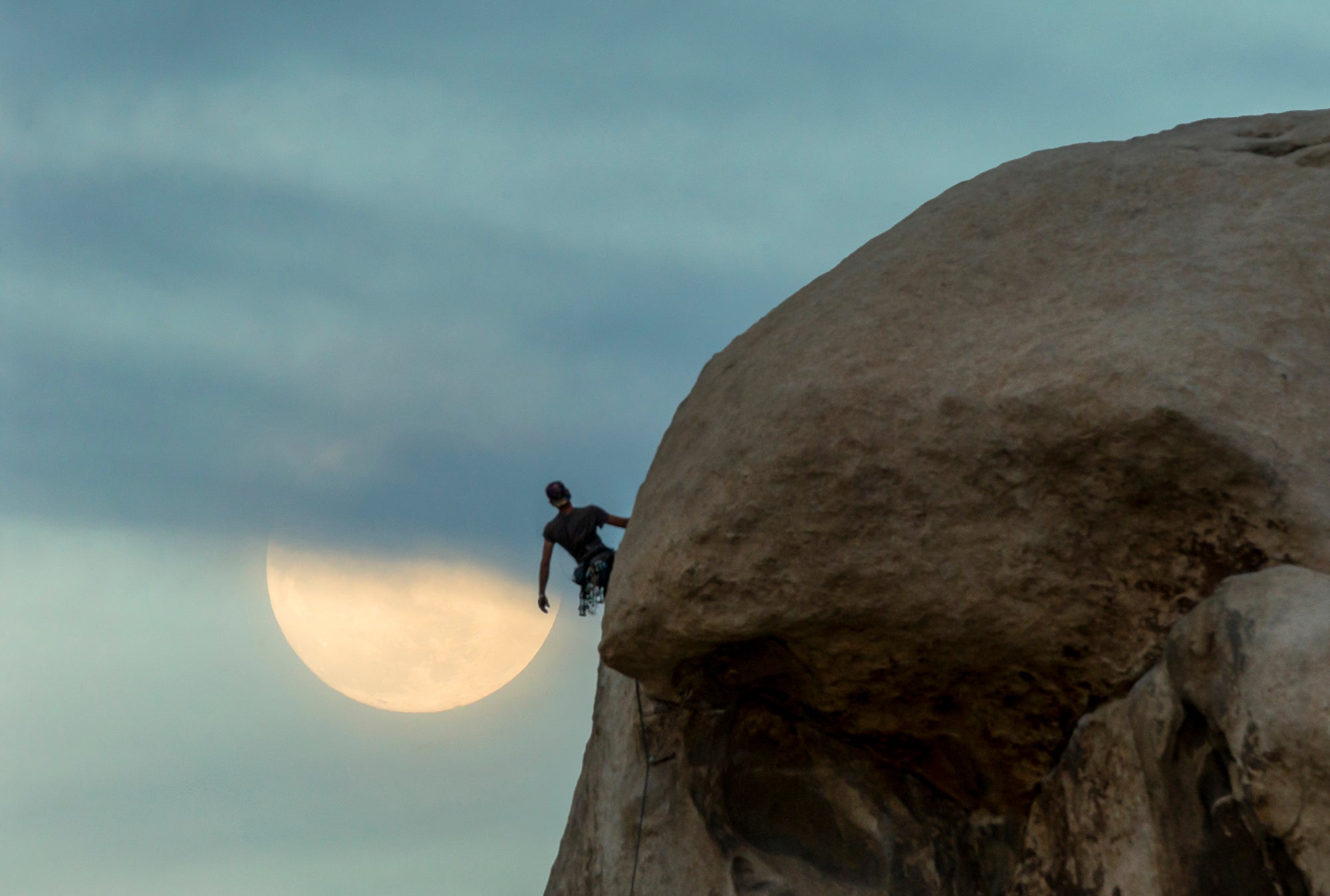 Rock climber on a sandstone dome at dusk with a large pale moon rising behind thin clouds.