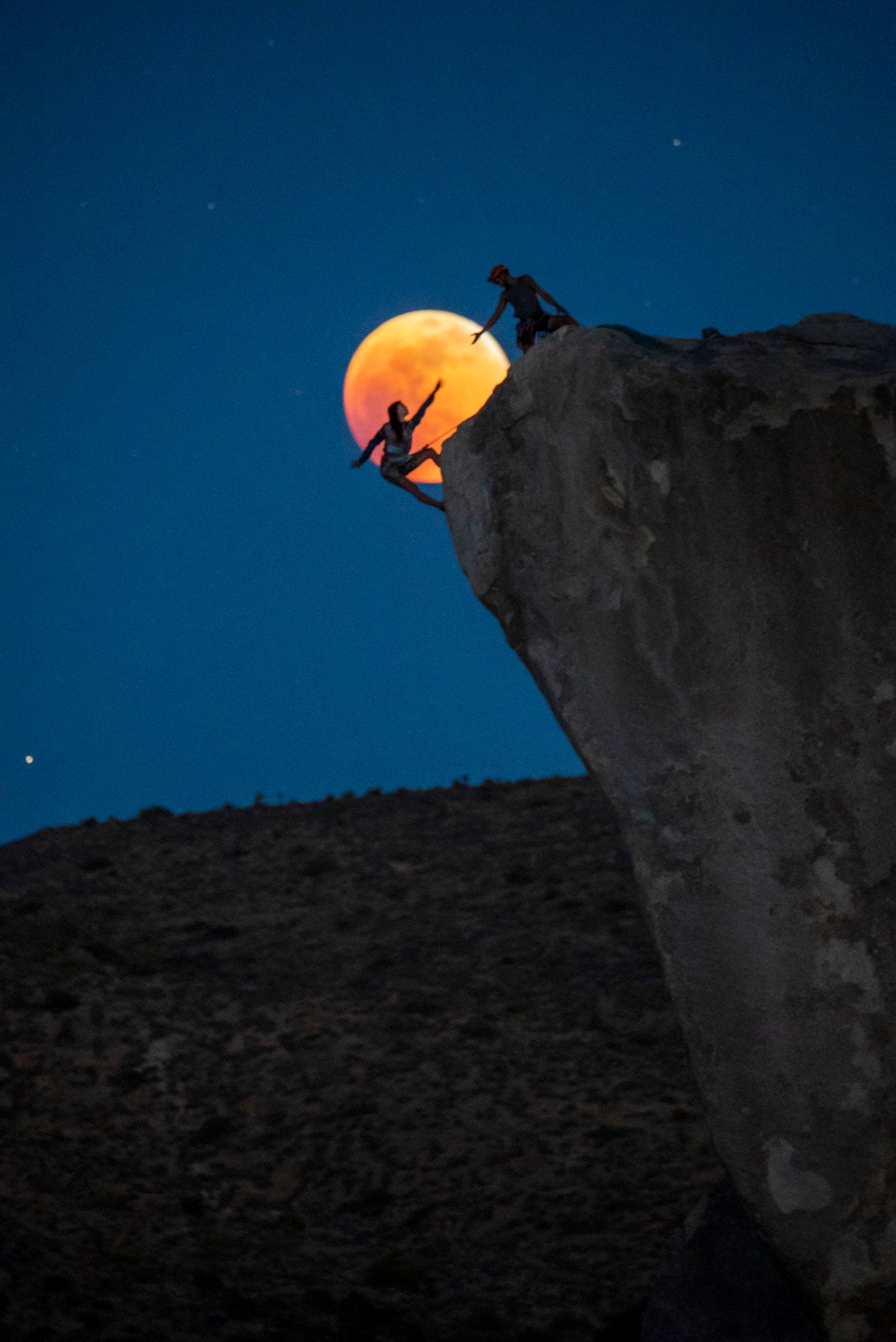 Two climbers silhouetted on a cliff edge as an orange-red eclipsed moon rises behind them.