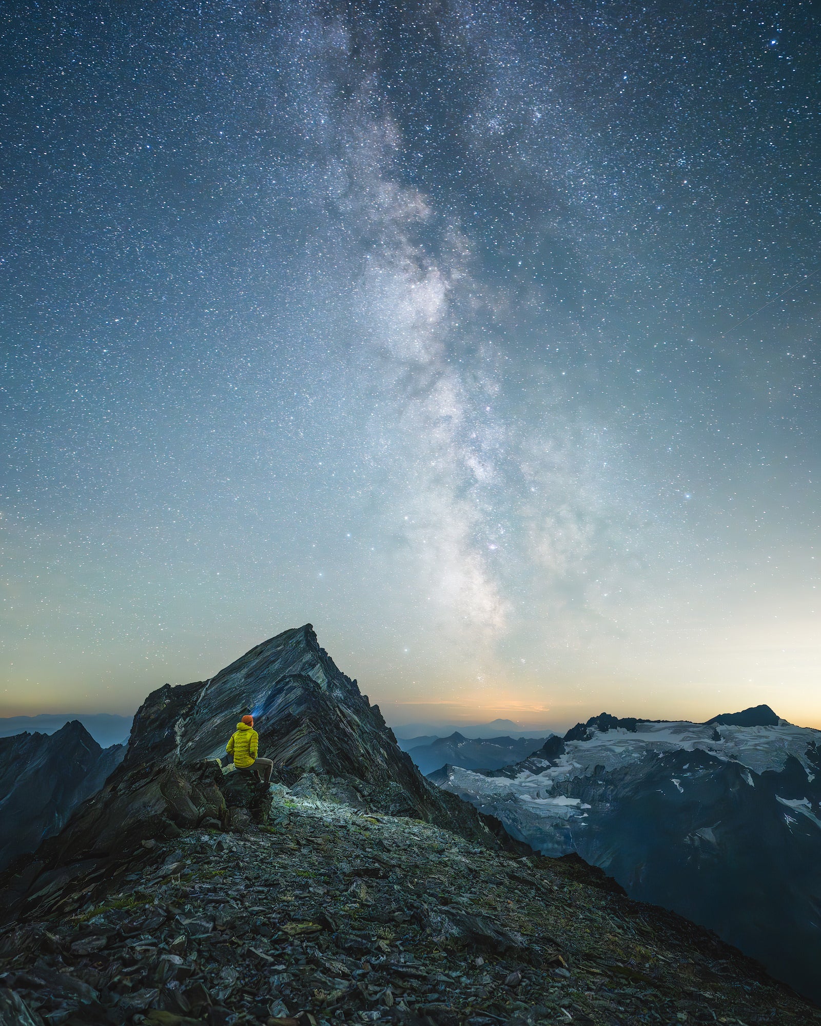 Milky Way rises above a jagged summit ridge as a seated figure looks out over glaciers and peaks in faint pre-dawn light.