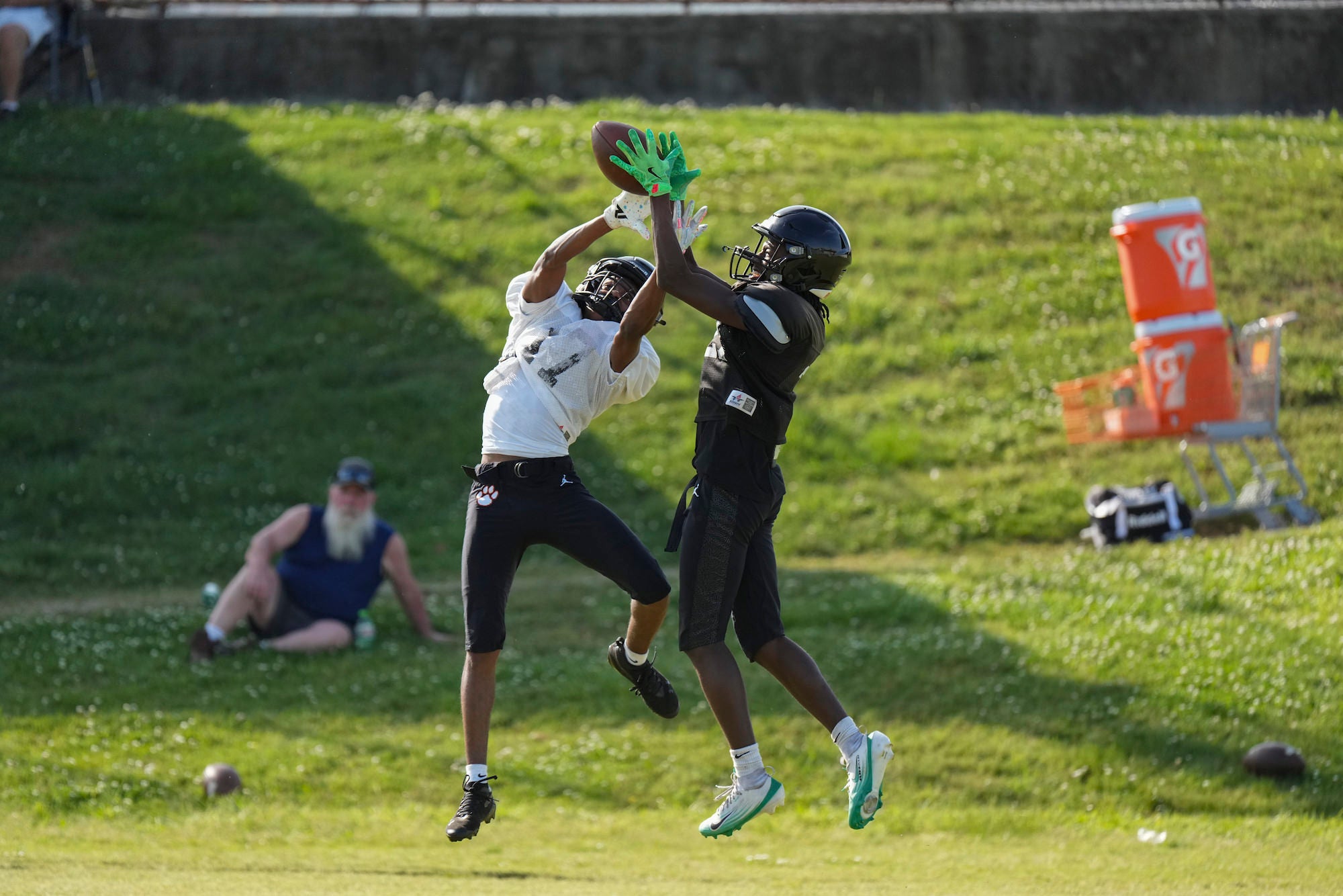 Two football players leap simultaneously to contest a pass, one in a white jersey and one in all black, both reaching upward with hands extended toward the football above them, on a sunlit green grass field during practice.