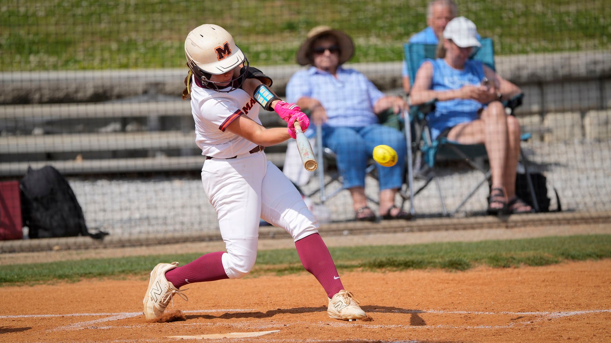 A softball player in a white and maroon Maryville uniform swings a bat at a yellow softball at home plate, with two spectators seated in folding chairs visible in the blurred background.