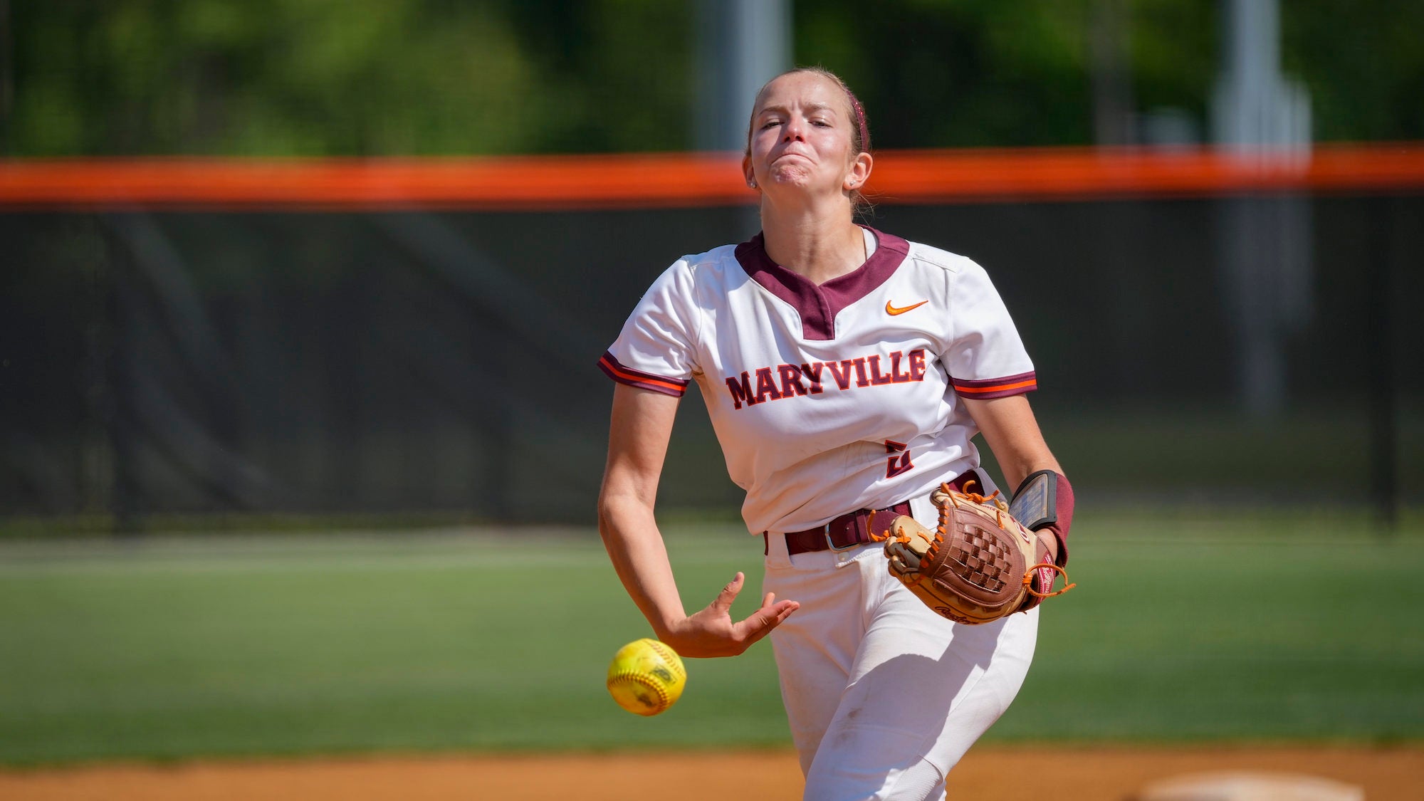A Maryville softball pitcher winds up to throw, holding a yellow softball at her side with a leather glove on the opposite hand, her expression focused and intense against a blurred green field and orange outfield wall.