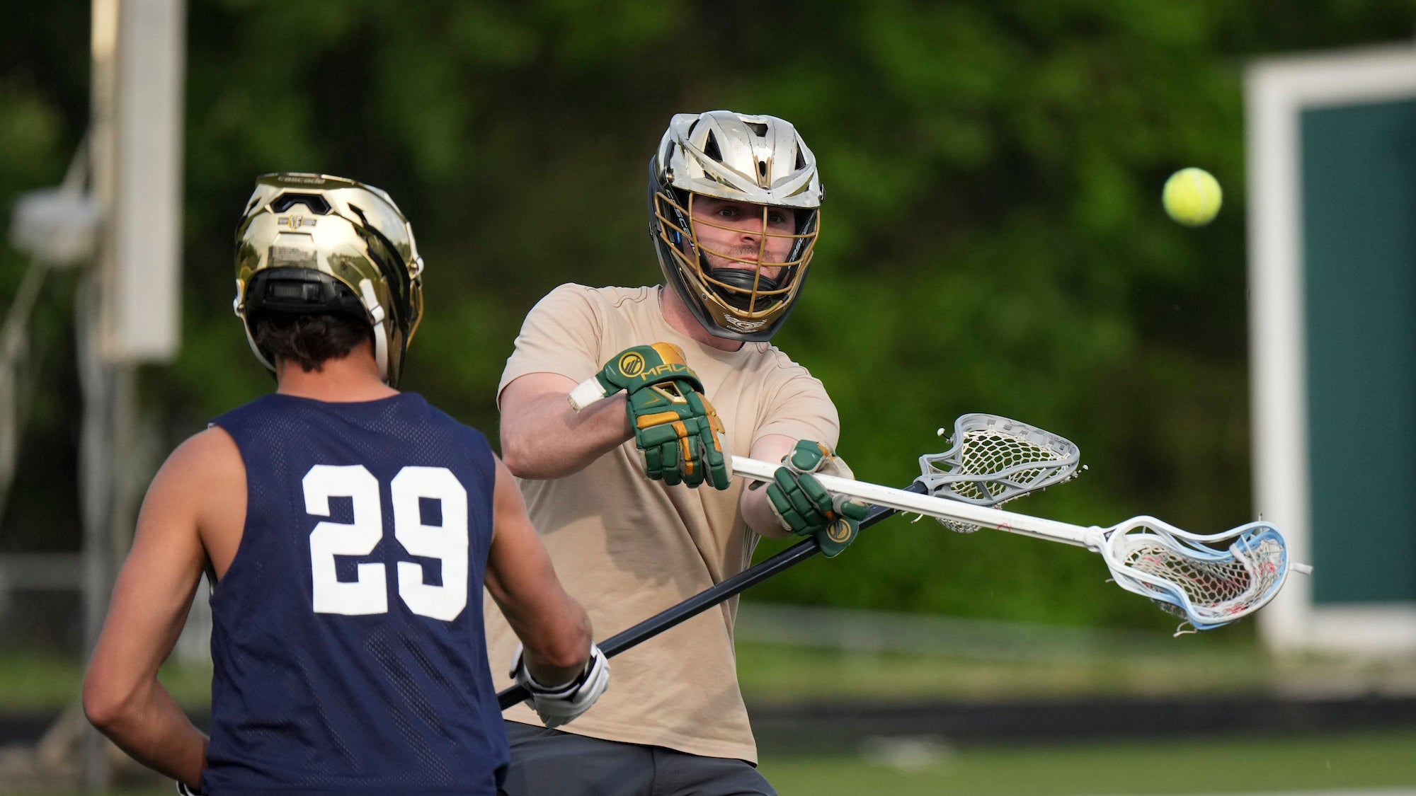 Two male lacrosse players face off during a game, with the player in the foreground wearing a white helmet and green-and-gold gloves cradling a lacrosse stick while a player in a navy number 29 jersey stands opposite him. A lacrosse ball is visible in the