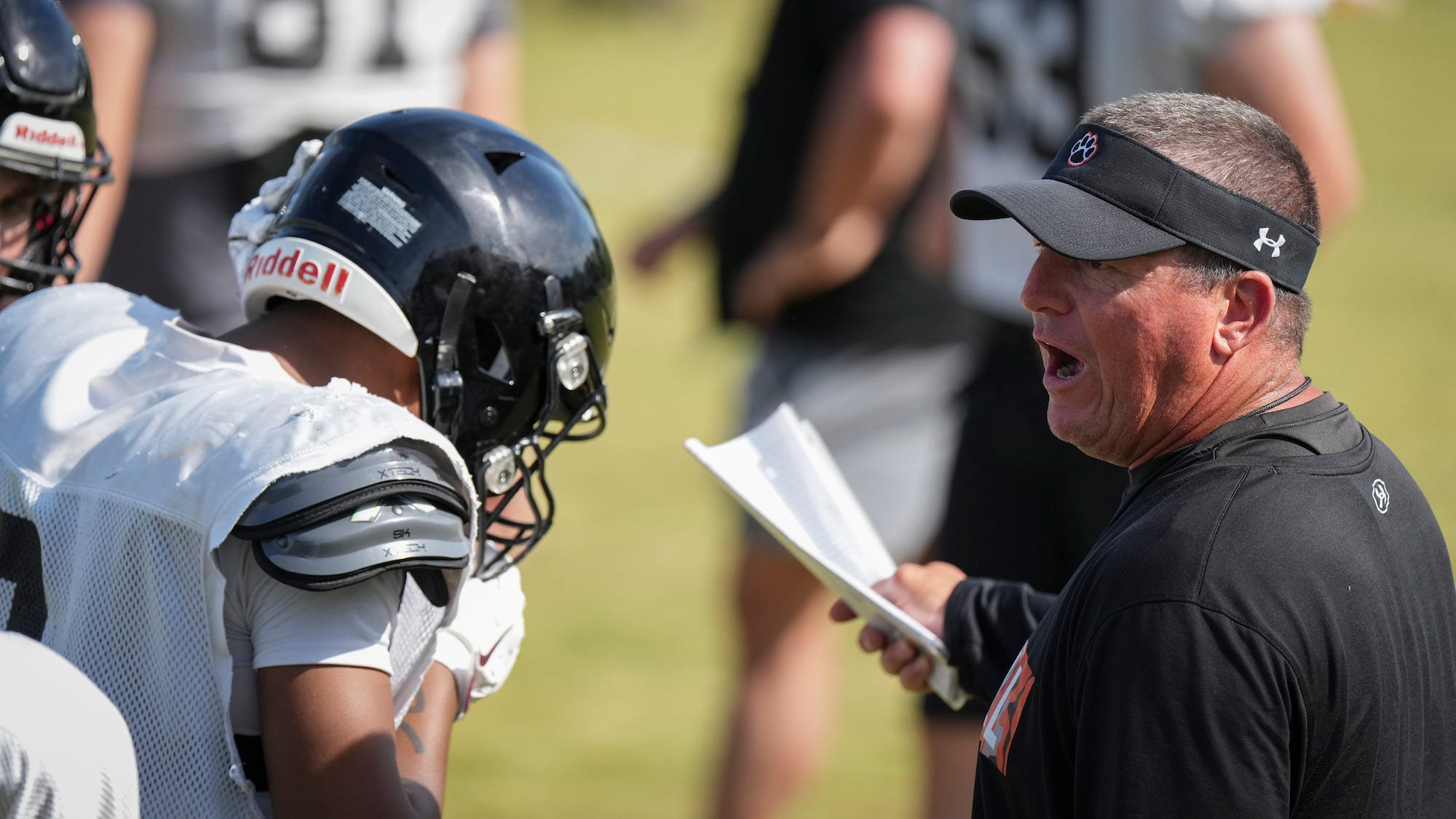Football coach holding papers talks to a football player.