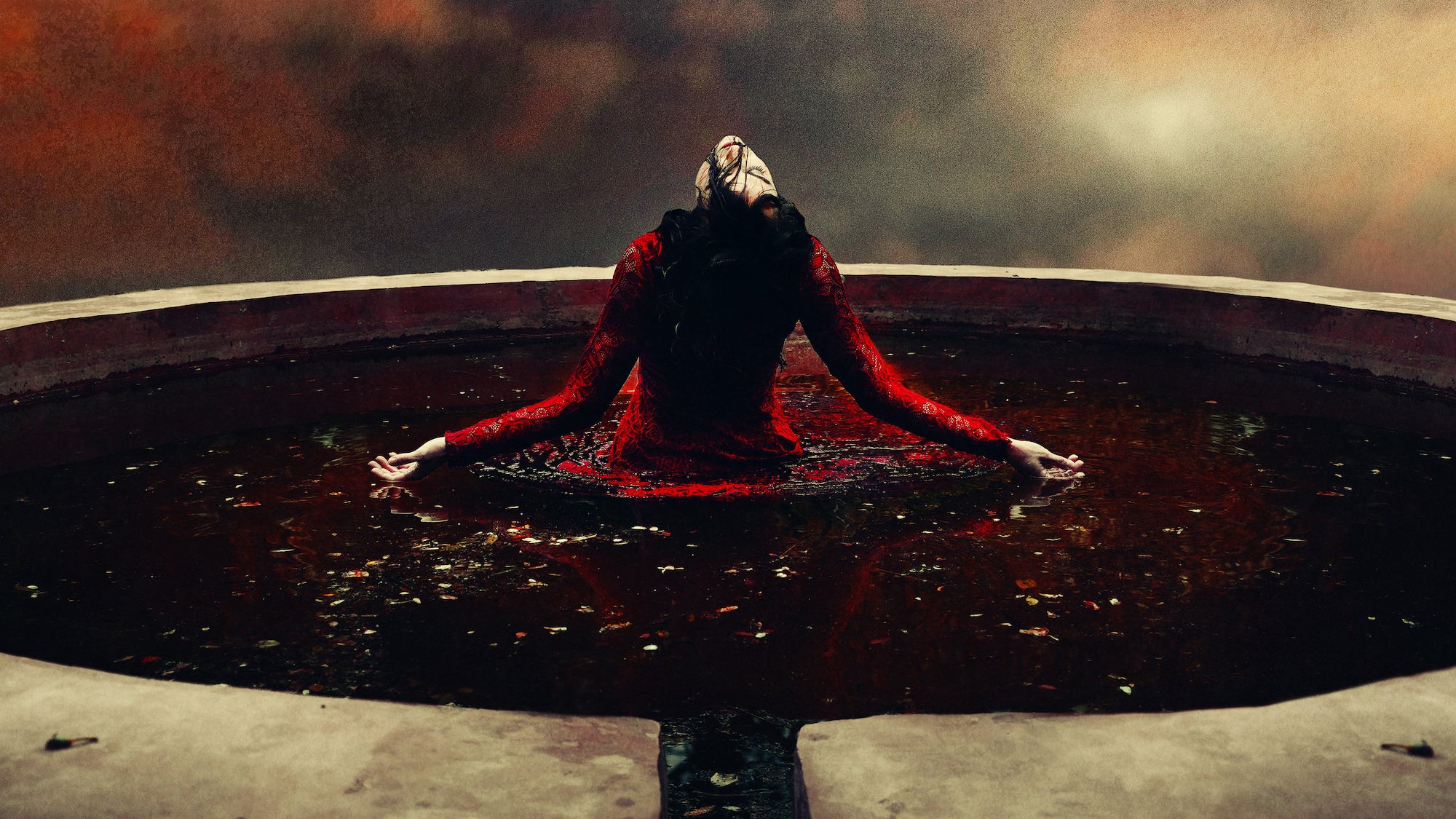 A wide-angle fine art image showing a masked figure in a red lace dress kneeling with arms outstretched in a circular stone fountain, surrounded by dark water and fallen leaves, beneath a moody, atmospheric sky.