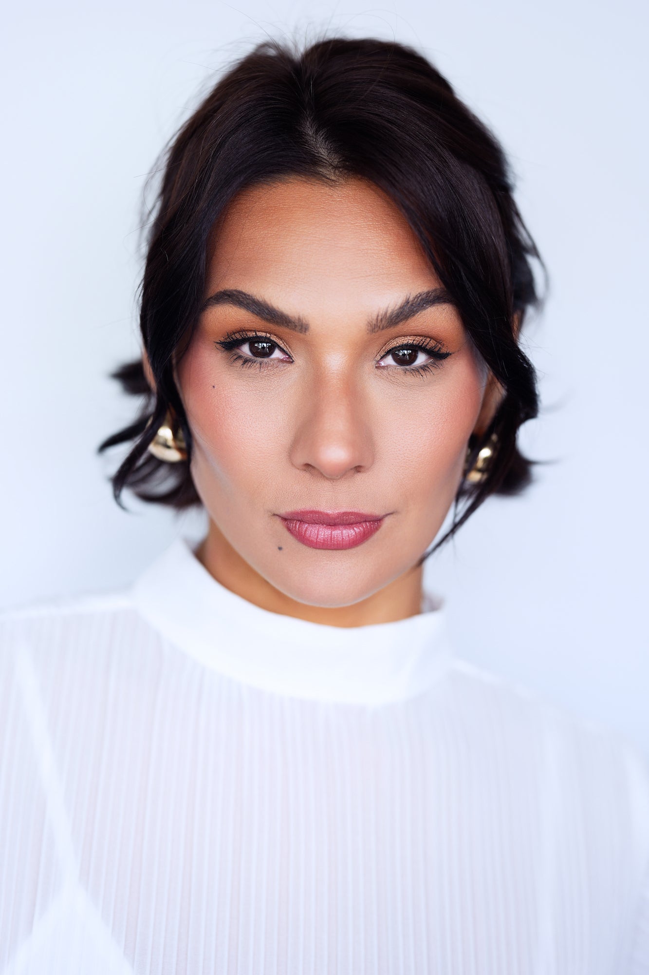 Studio-style portrait of a woman with dark hair pulled back, wearing a white ribbed mock-neck top and gold earrings, gazing directly into the camera against a clean light grey background.