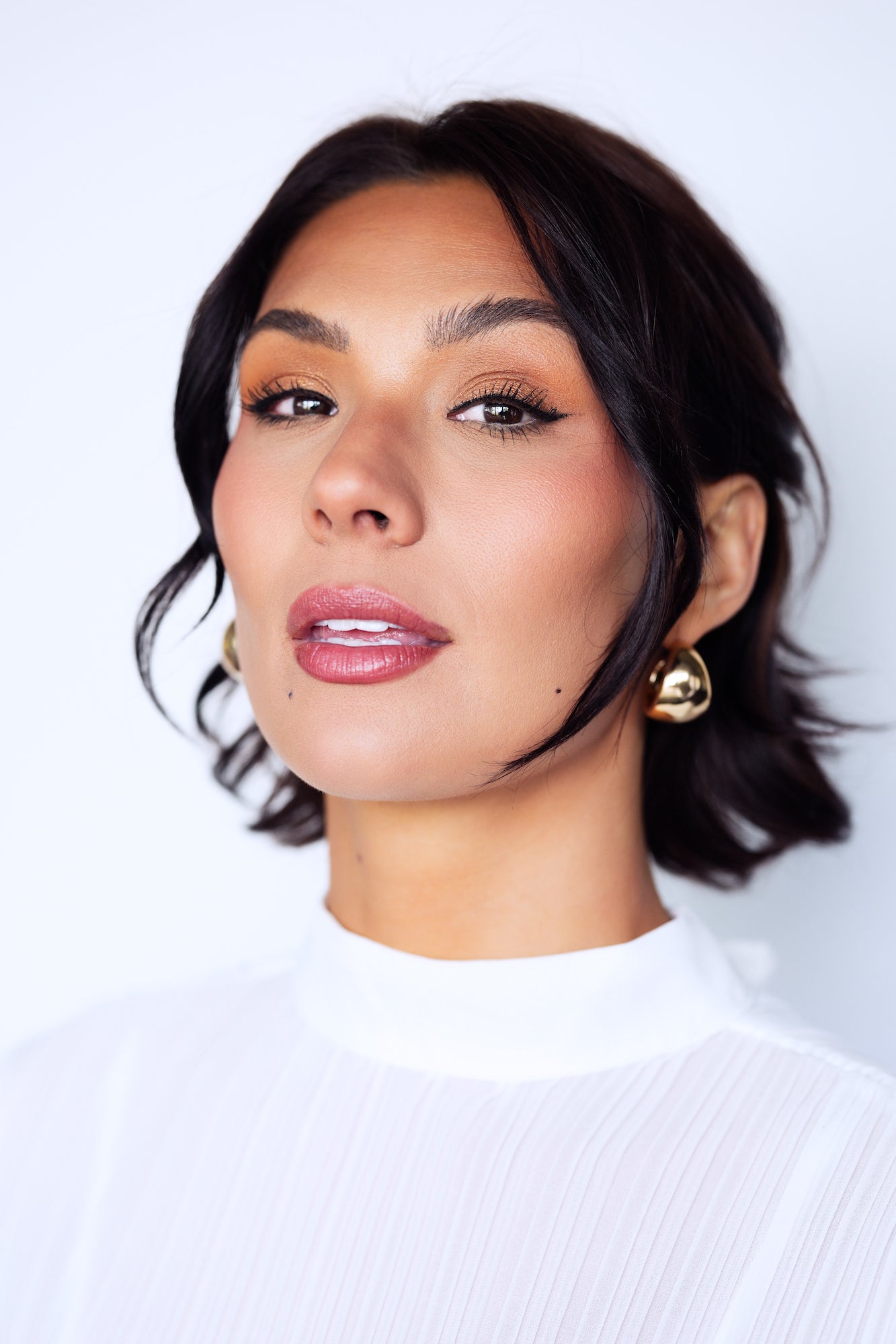 Beauty portrait of a woman with dark wavy hair, gold sculptural earrings, and pink lips wearing a white ribbed top, shot against a clean light blue-grey background with a soft, slightly parted-lip expression.