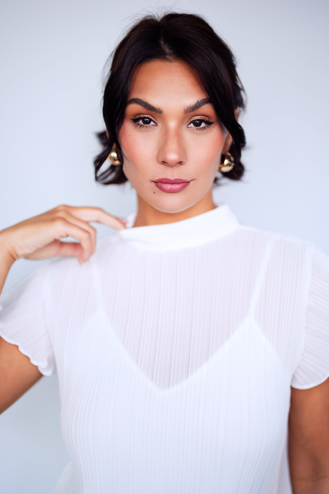 Close-up beauty portrait of a woman with dark wavy hair and gold earrings, wearing a white ribbed top, looking directly into the camera against a soft light grey background with a warm, confident expression.
