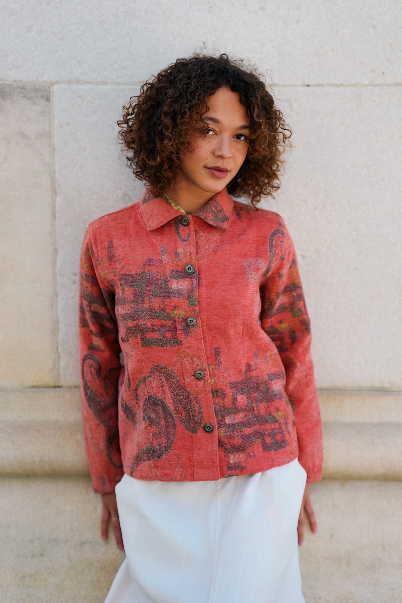 Full-length portrait of a young woman with curly hair standing against a stone wall, wearing a red paisley button-up jacket over white trousers, looking off to the side with a relaxed, natural expression.