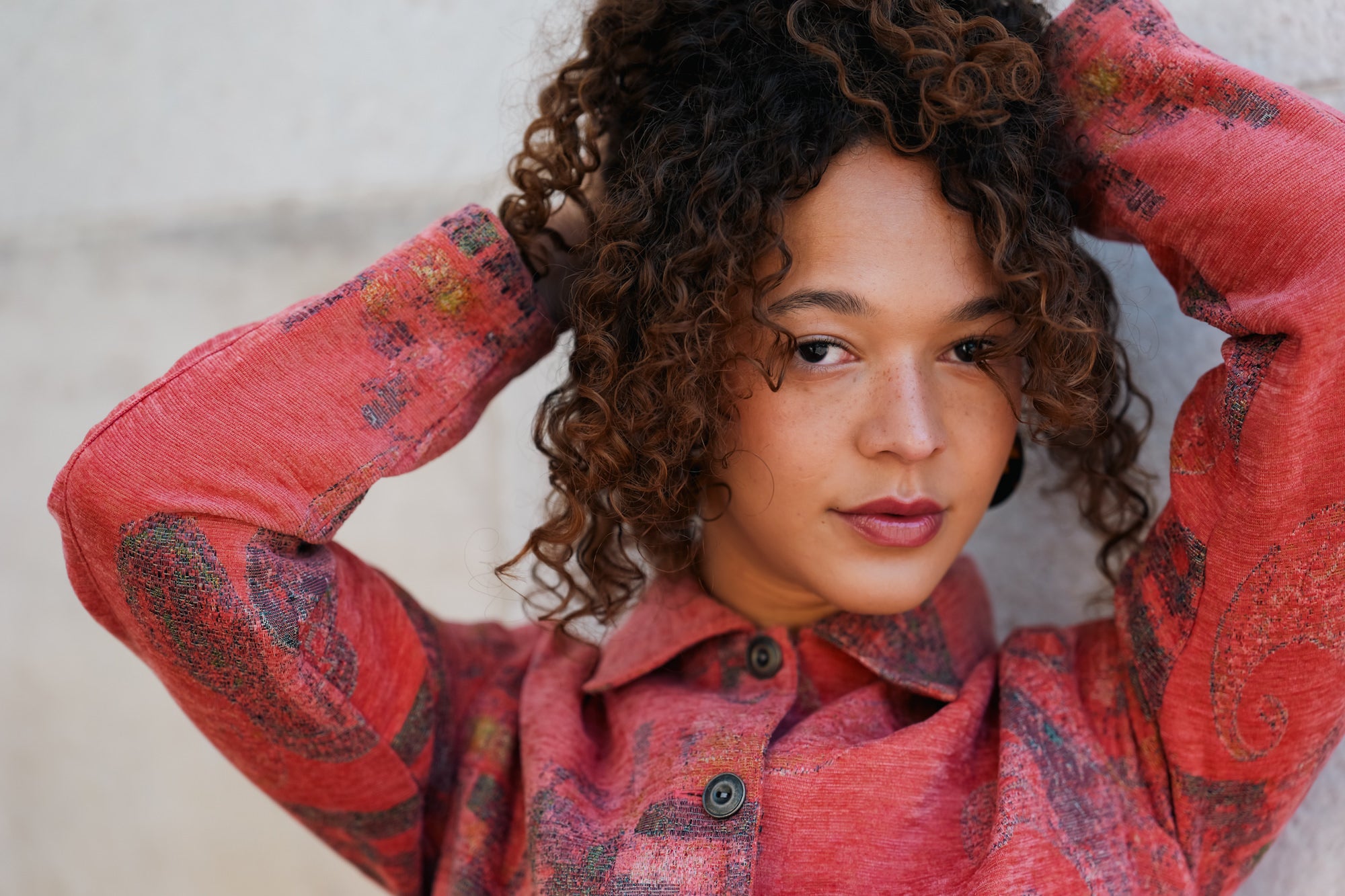 Close-up portrait of a young woman with voluminous curly brown hair, wearing a red patterned jacket, her hands raised behind her head as she gazes off to the side.