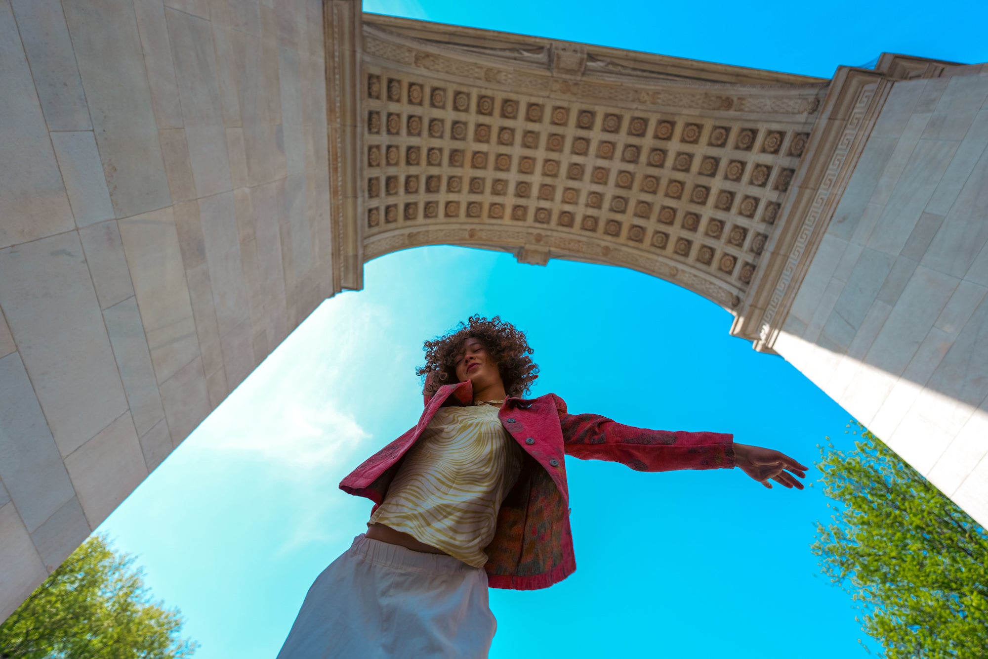 Dynamic low-angle portrait of a young woman with curly hair spreading her arms wide beneath the ornate coffered arch of a grand monument, shot from below against a bright blue sky with green trees framing the edges.