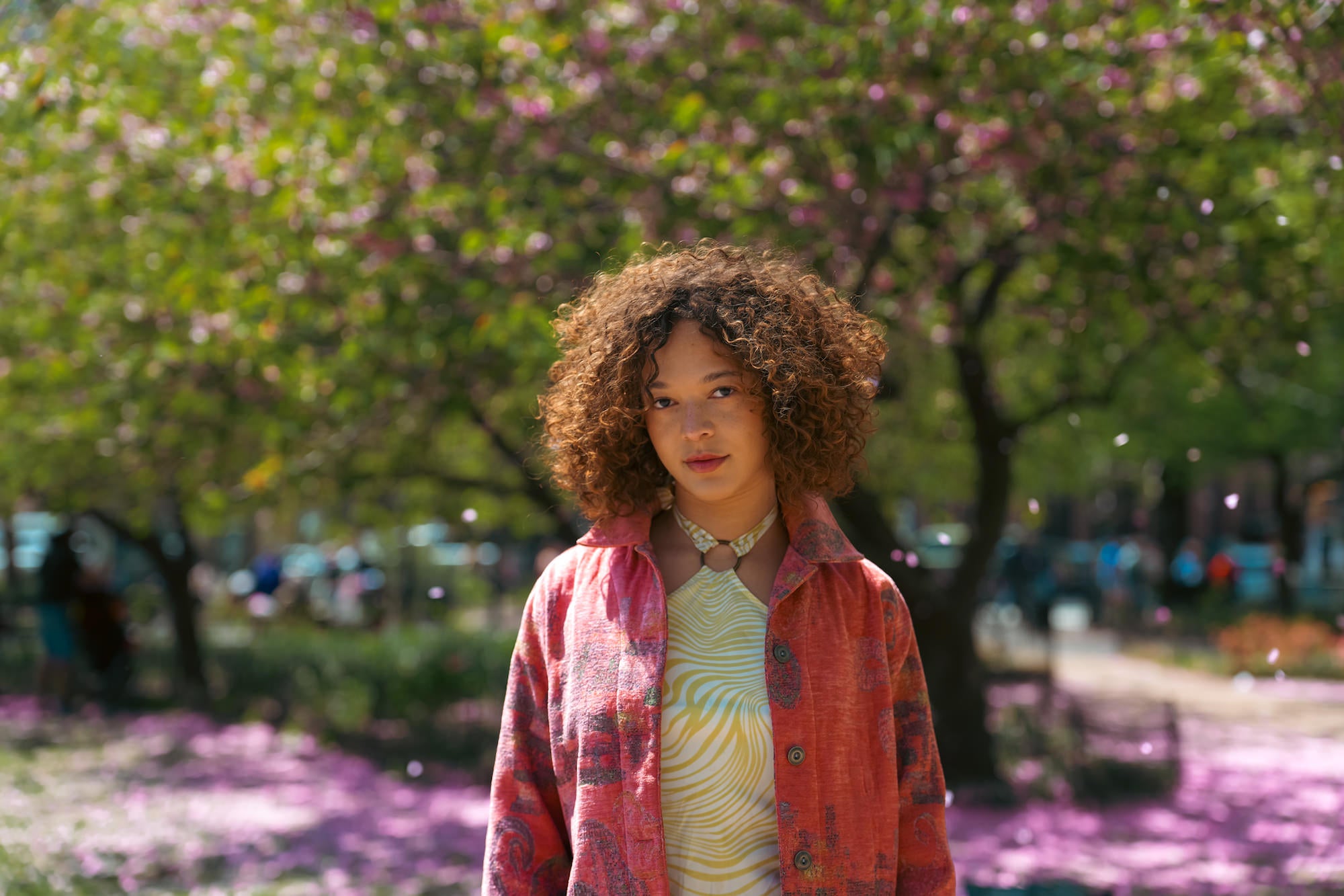 Outdoor portrait of a young woman with curly honey-brown hair standing in a sunny park lined with flowering trees, wearing a red floral jacket over a yellow swirl-print top with a ring choker necklace, surrounded by scattered pink petals on the ground.