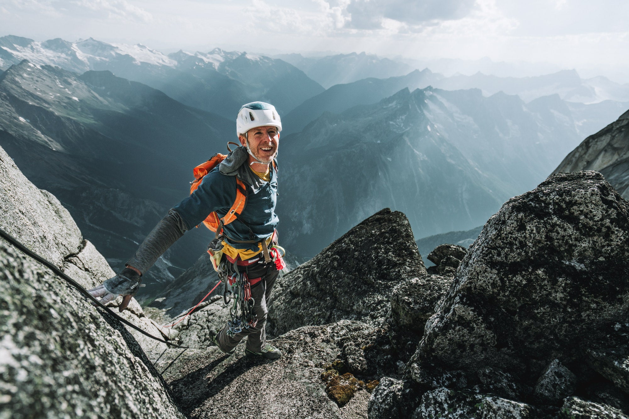 Climber Tommy Caldwell in climbing gear standing on a mountain with more mountains in the background.