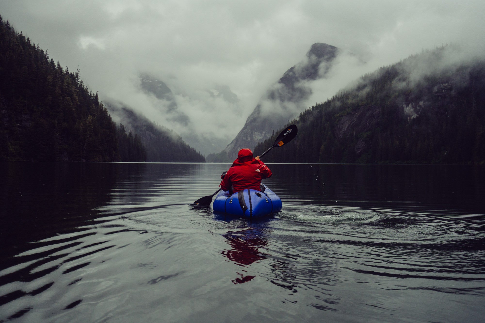 Man in red coat kayaking in lake with mountains in the background.