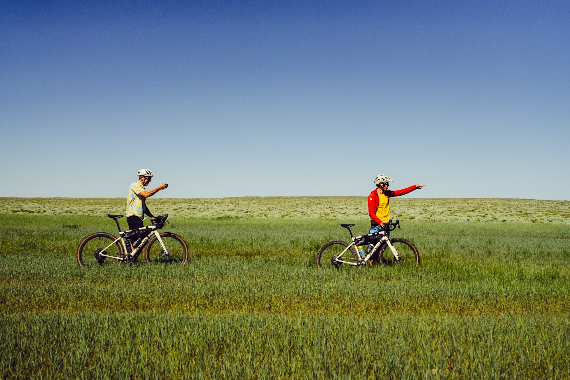 Alex Honnold and Tommy Caldwell in a green field with their bikes, both pointing with their hands forward to the right.