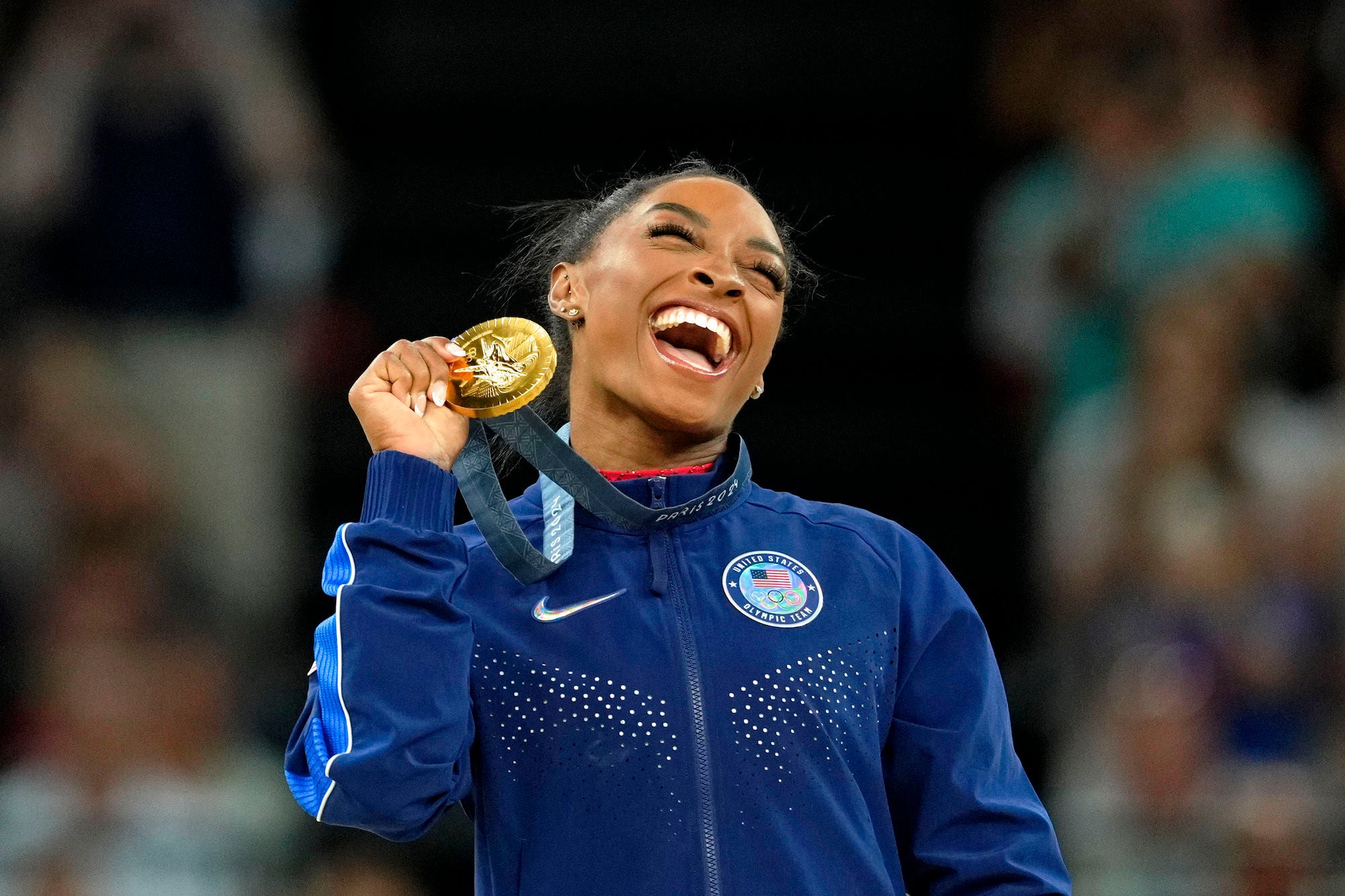 Gymnast smiles and displays a gold medal while wearing a Team USA jacket.