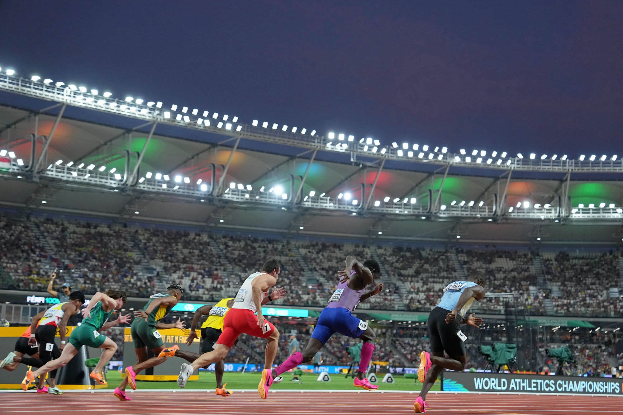 Sprinters explode from the blocks in a packed stadium at dusk.