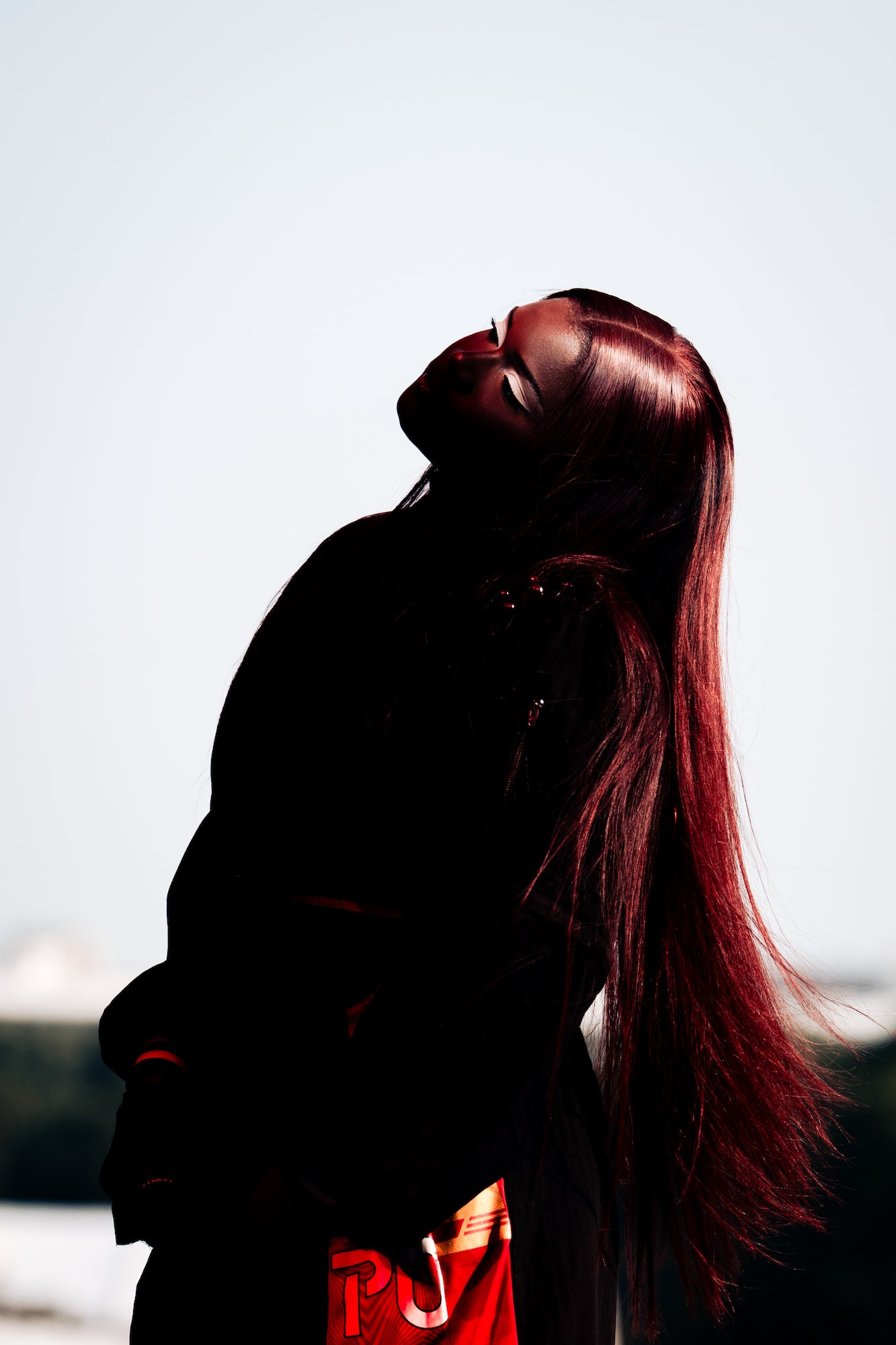 Backlit portrait with long auburn hair; head tilted, dramatic contrast against sky.