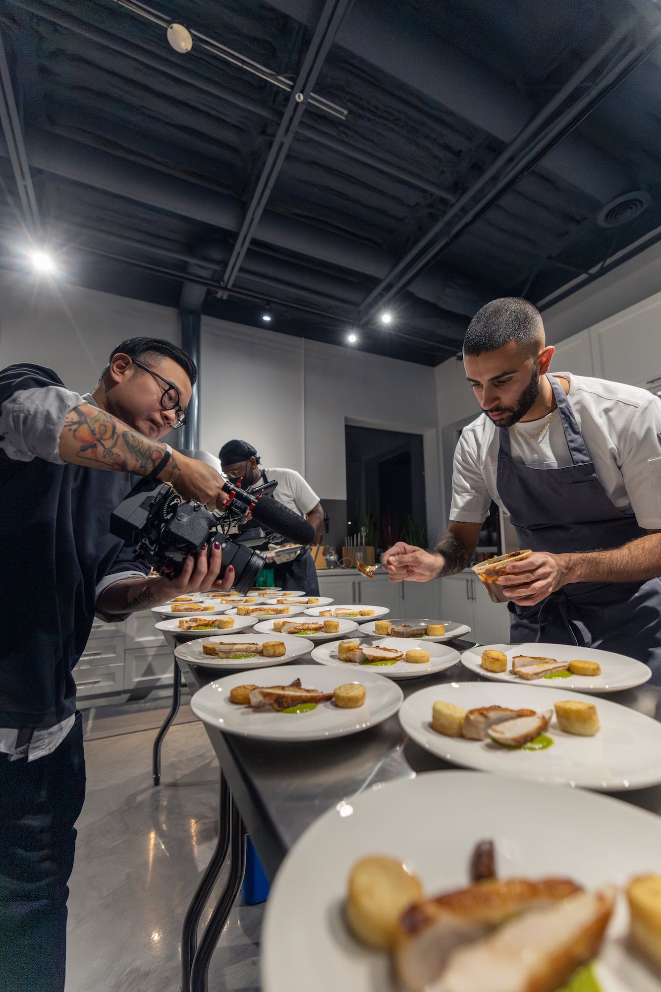 Videographer films a chef plating multiple identical dishes on a stainless table in a pro kitchen.