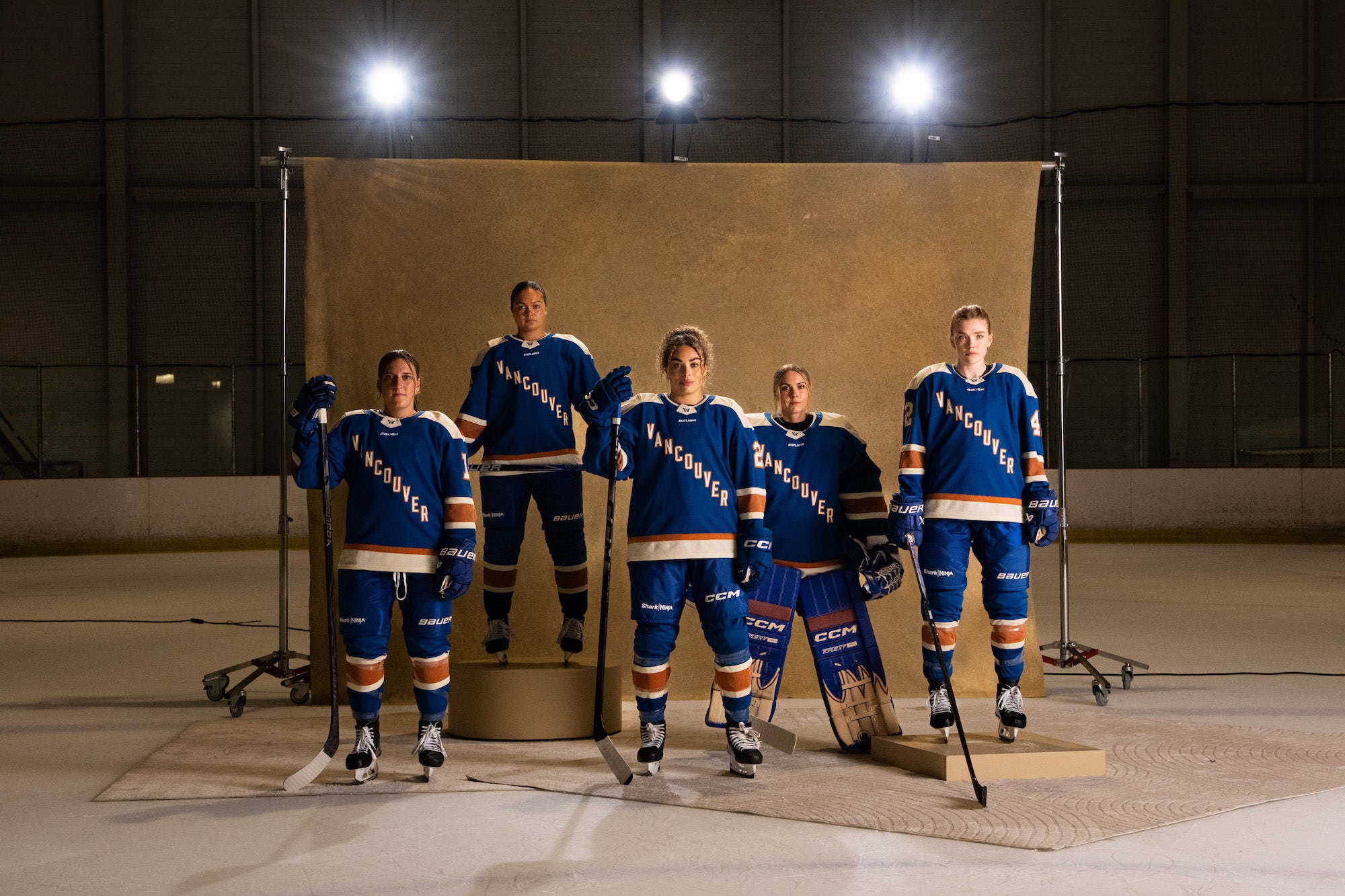 Five players in blue &ldquo;Vancouver&rdquo; uniforms pose on ice in front of a studio backdrop and lights.
