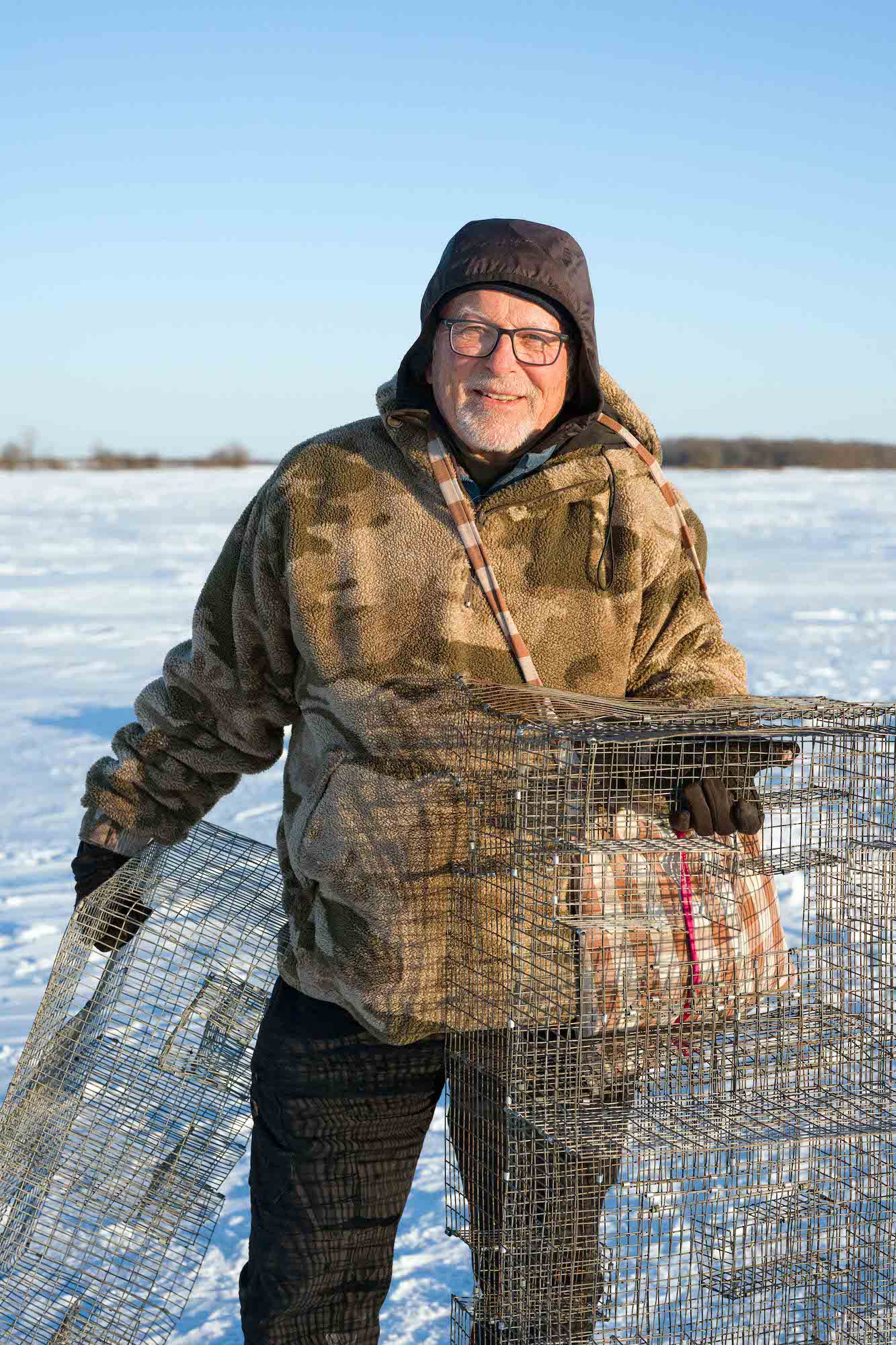 Person in heavy winter clothing smiling while holding wire cages on a snowy field. Person in heavy winter clothing smiling while holding wire cages on a snowy field.