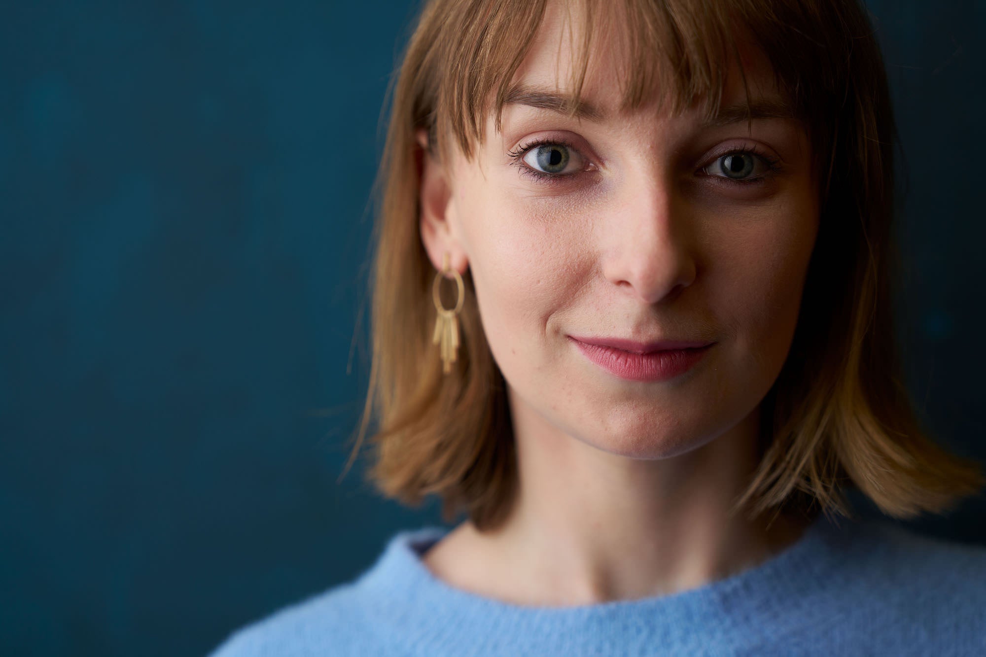 Close-up portrait of a woman in a blue sweater with gold earring, teal background and shallow depth of field.