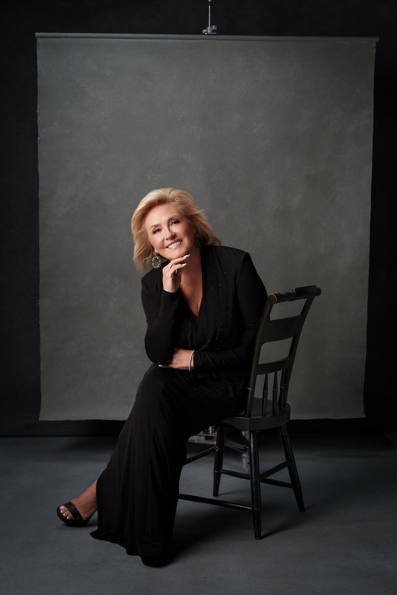 Studio portrait of a woman in a black outfit seated on a chair, smiling with chin rested on hand against a gray backdrop.
