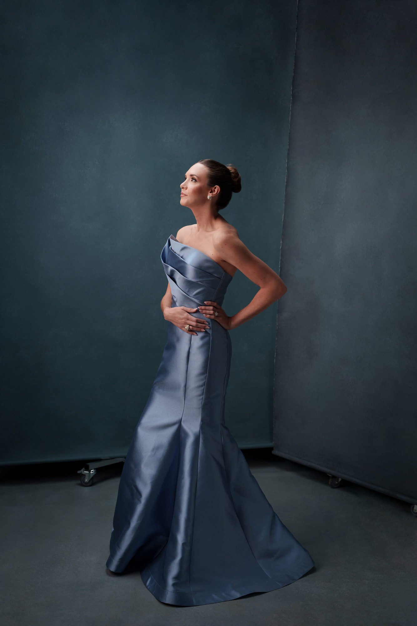 Woman in a strapless slate‑blue evening gown posing in profile with hands on hips against a textured studio backdrop.