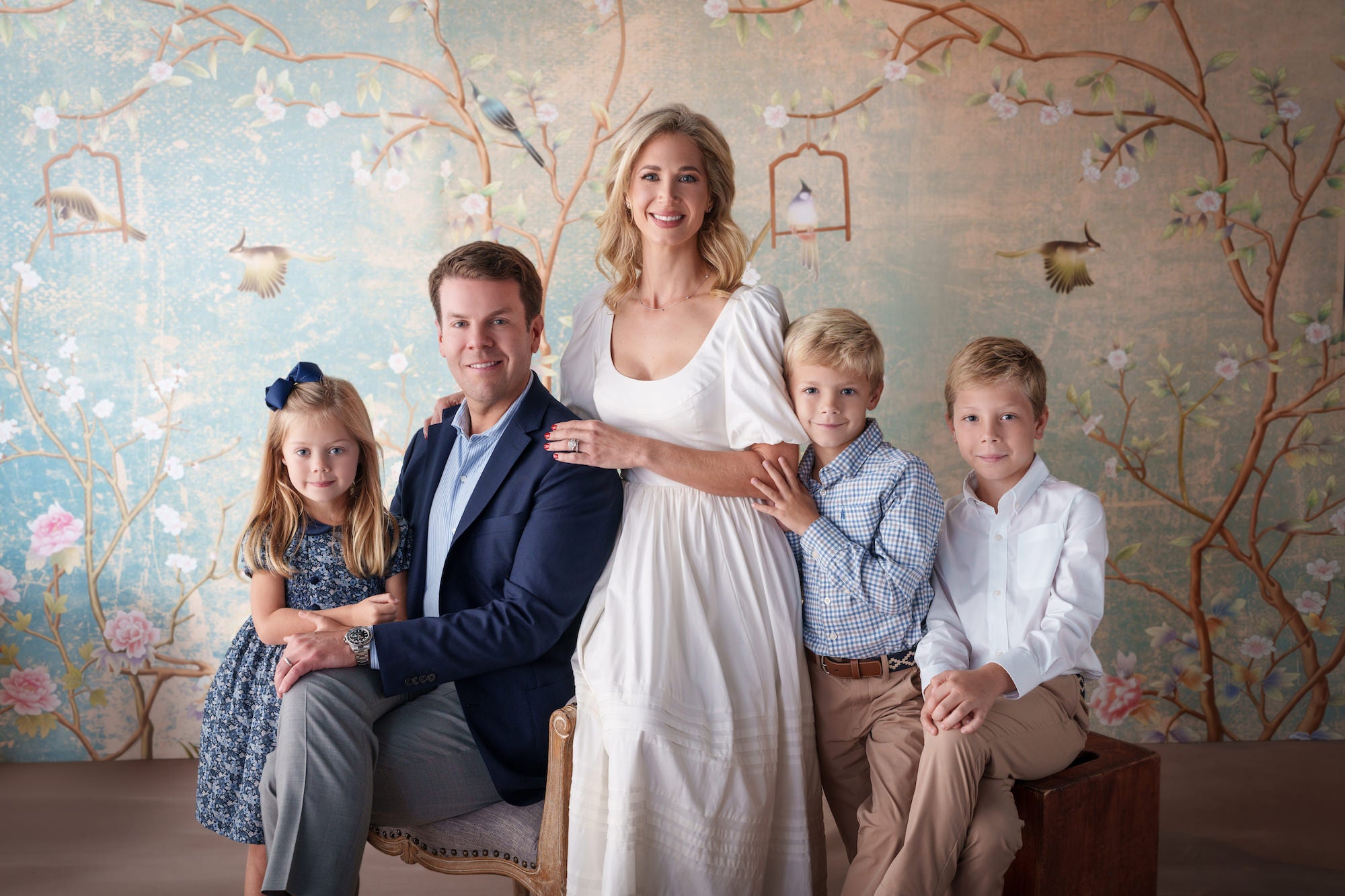 Studio family portrait of five in soft blues and whites, posing in front of a pastel bird‑and‑floral mural backdrop.