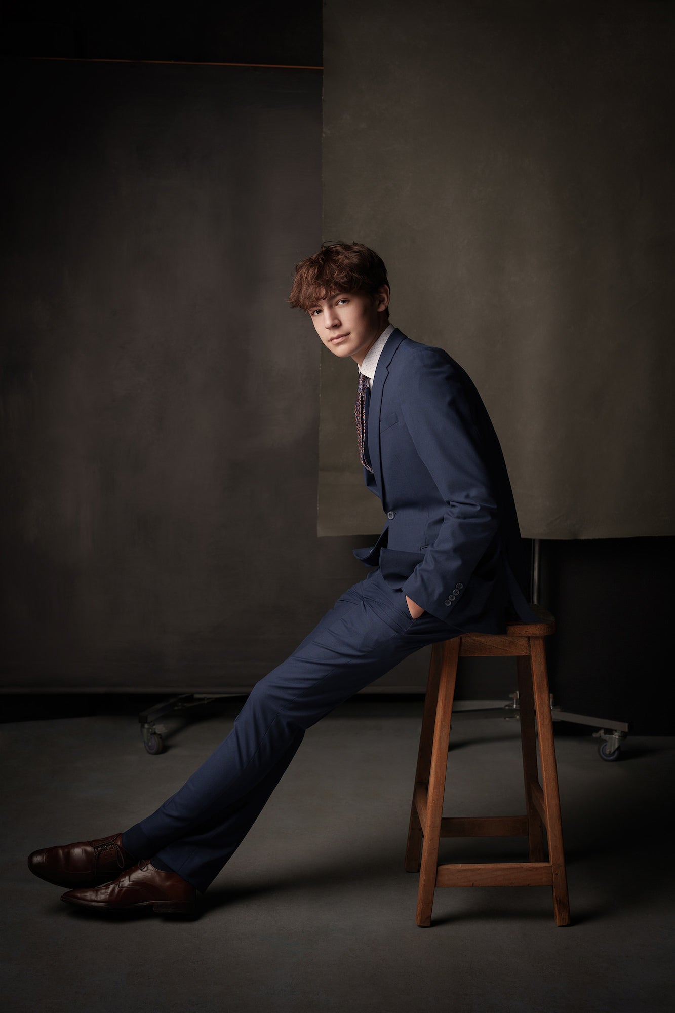 Young man in a blue suit seated on a wooden stool in a studio, legs extended and hands in pockets, moody backdrop.
