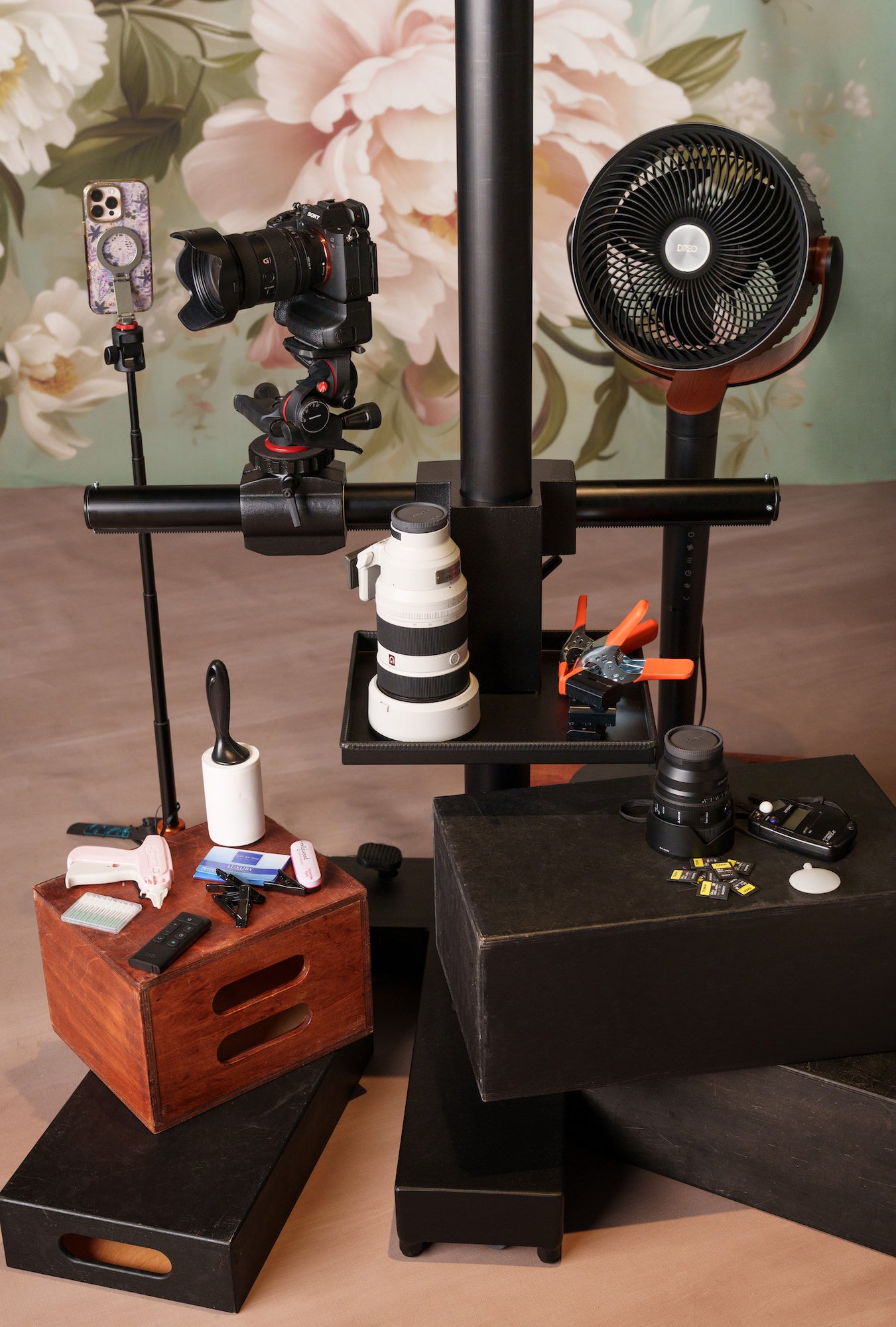Studio gear setup with mirrorless camera on stand, telephoto and prime lenses, phone on clamp, fan, memory cards, and accessories.